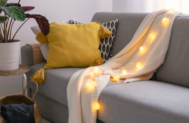 The 2021 Pantone Color of the Year is gray and yellow. In this image, a yellow pillow and decorative lighted throw are on a gray sofa.